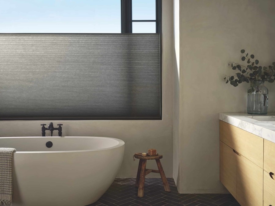 Modern bathroom featuring a large window with Hunter Douglas cellular shades in a dark gray color, filtering natural light above a freestanding oval bathtub.
