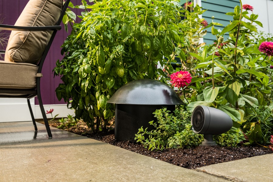 A Klipsch outdoor speaker and subwoofer nestled among foliage, flowers, and shrubs.