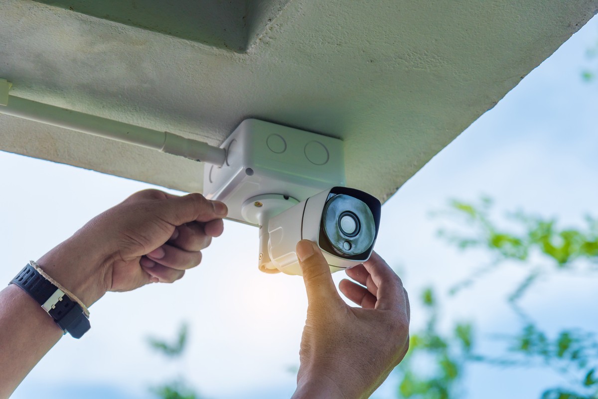 A man’s hand is adjusting a security camera mounted under the eave of a roof. 